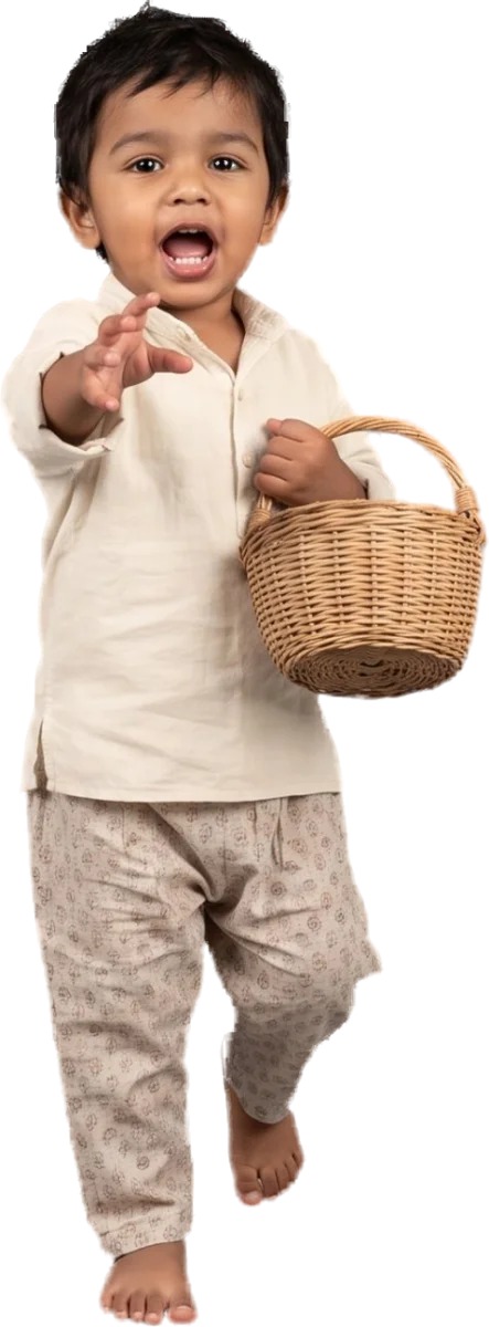 A toddler in a cream shirt and printed trousers walking towards the camera, mid-sentence, reaching out with one hand and holding a small woven basket.