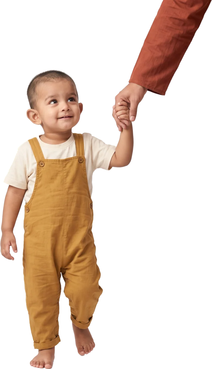A toddler in mustard dungarees holding an adult’s hand and looking up with a small smile.
